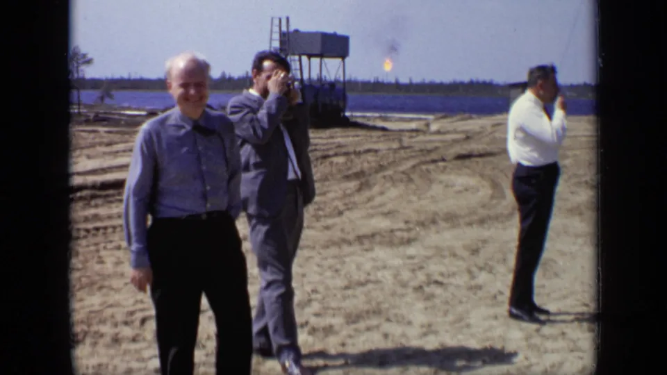 1971 Moscow construction-site frame showing men in shirts and suits near dirt tracks, water, and a small distant flare or flame.
