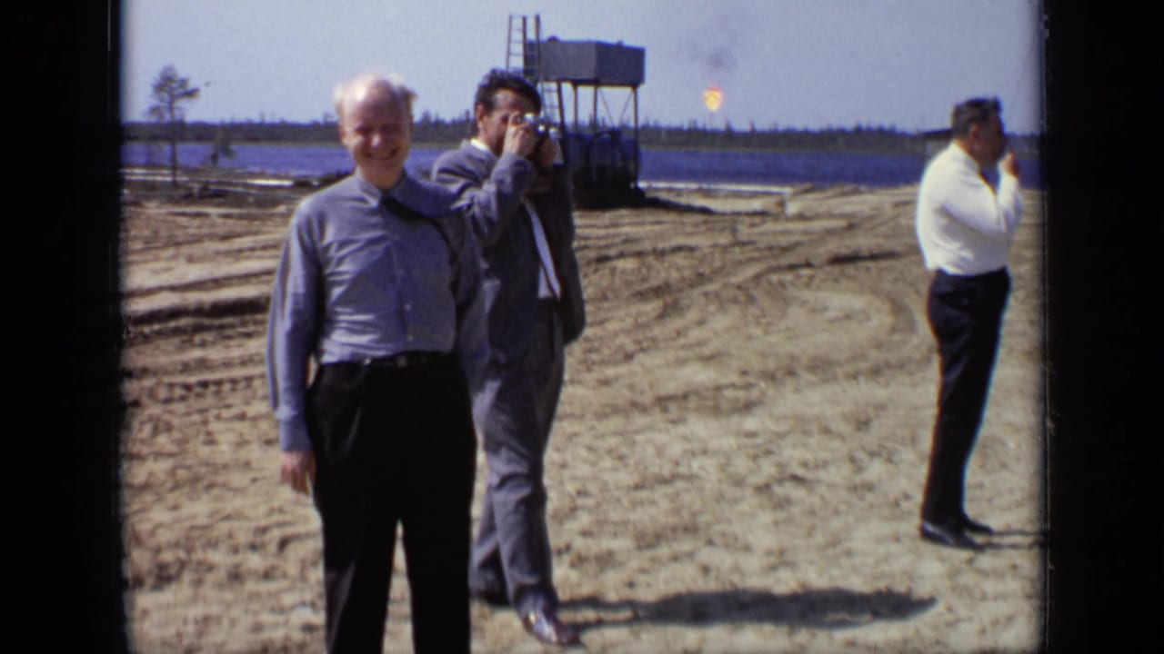 Men in suits stand on a cleared sandy construction site, with one man raising a camera near industrial structures.