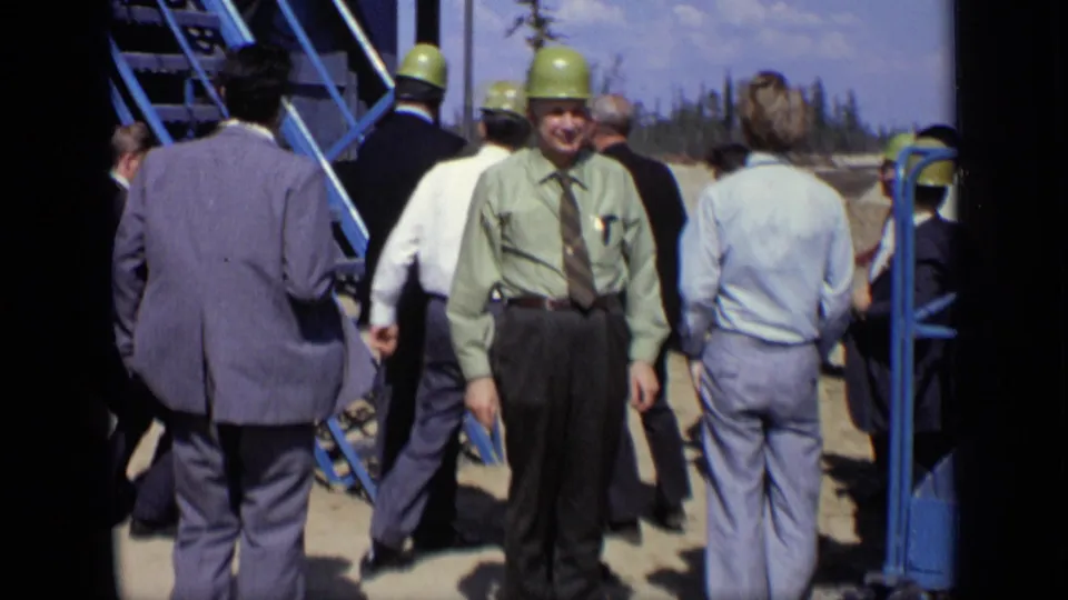 1971 Moscow frame of adult men in suits and green hard hats gathered near a blue metal staircase at a cleared industrial site.