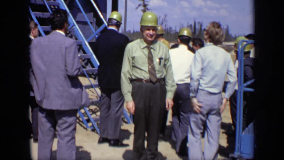 1971 Moscow home-movie frame showing a smiling hard-hatted man facing the camera while others stand near a blue staircase.