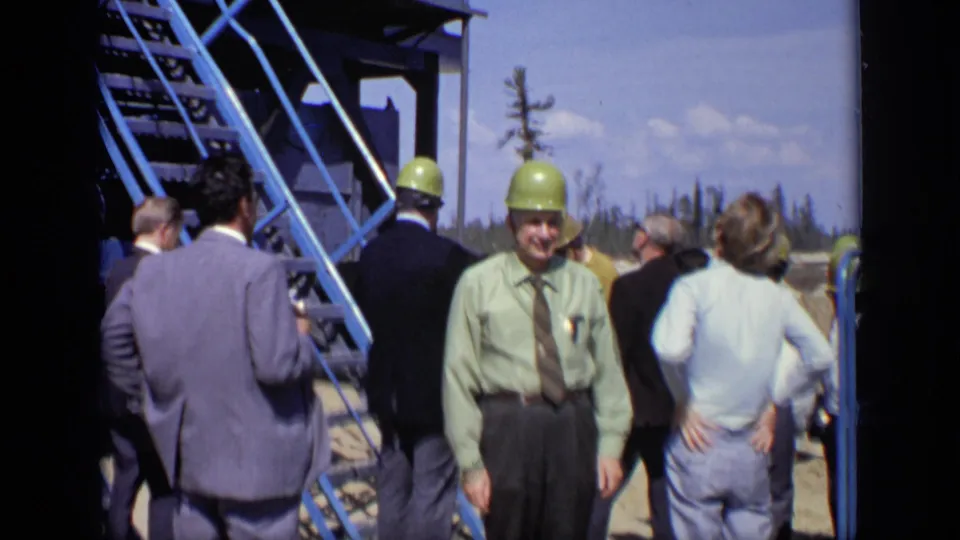 1971 Moscow frame with suited adult men and green hard hats clustered beside a blue metal staircase at an industrial construction site.