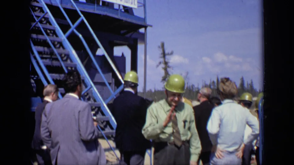 1971 Moscow footage shows suited men and hard-hatted visitors gathered beside a blue industrial stairway with a Cyrillic sign overhead.
