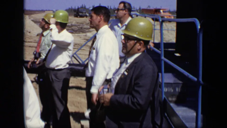Several men in hard hats, shirts, and suits look across a sandy industrial construction area in 1971 Moscow home-movie footage.