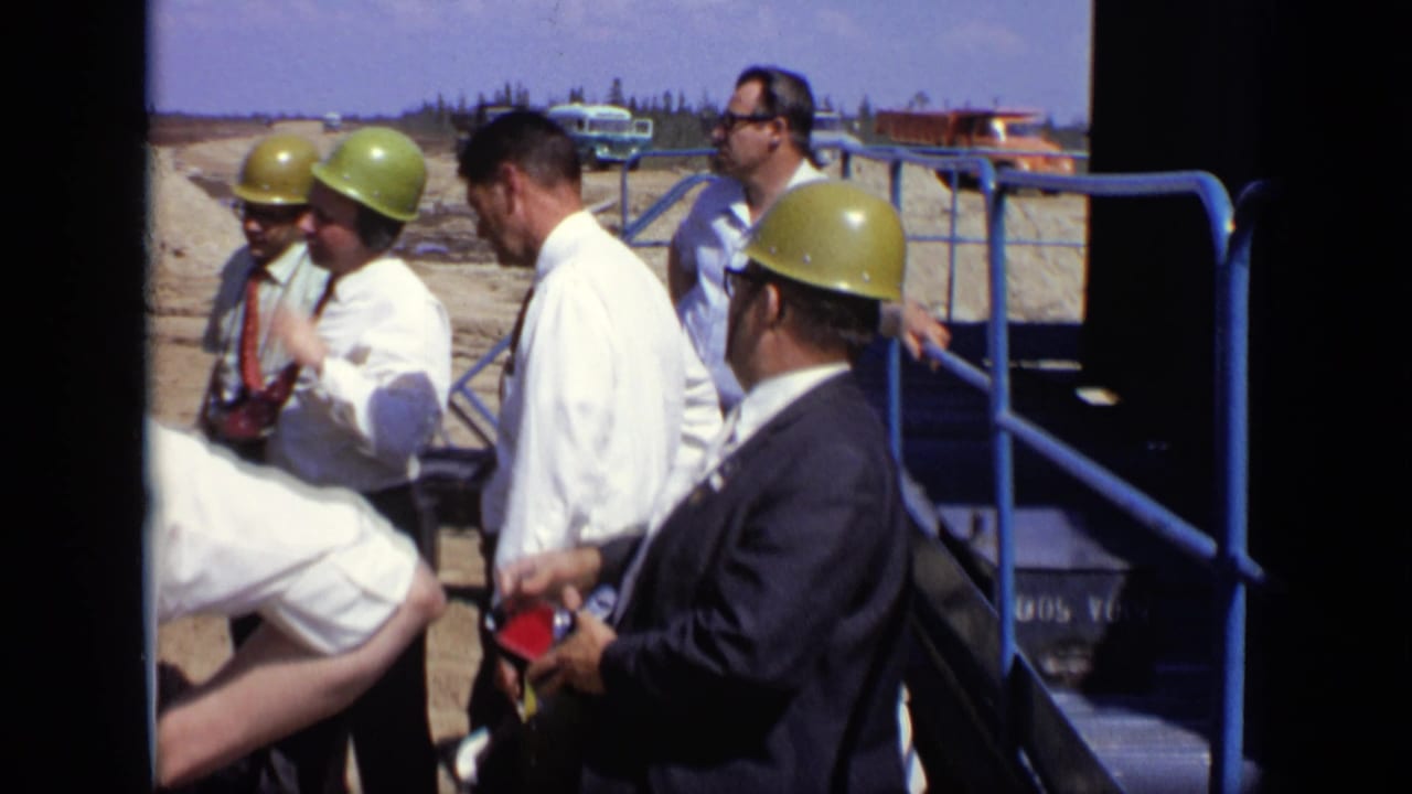 Men in yellow hard hats and dress shirts stand near blue railings at an outdoor construction site.
