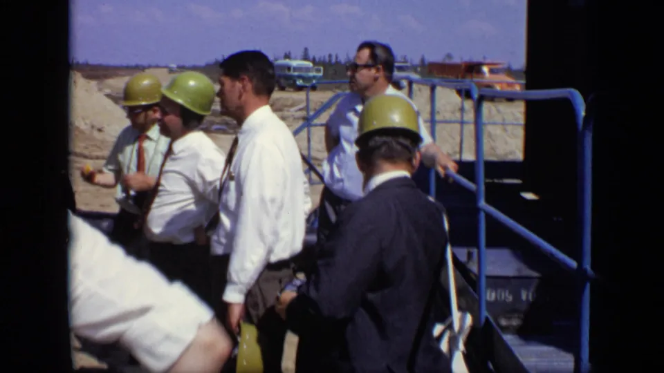 The 8mm frame shows a small hard-hatted group in business attire gathered at a Moscow work site, with blue railings and vehicles behind them.