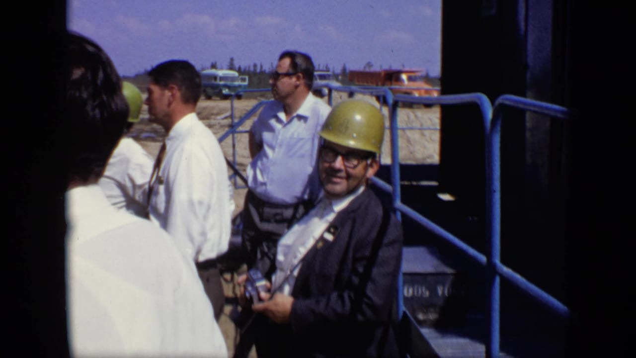 1971 Moscow construction-site visitors stand near blue railings, including a suited man in a yellow hard hat looking toward the camera.