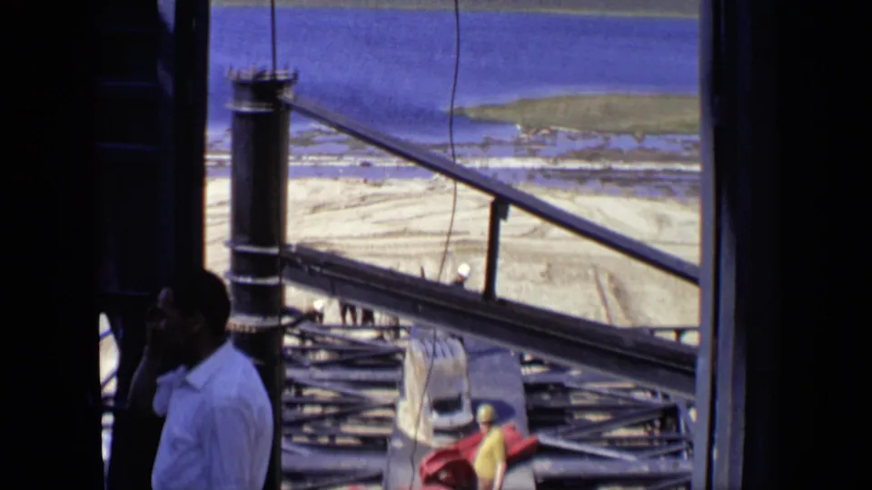 The camera looks past men and railings across a construction platform in Moscow, showing steel framing, sandy earth, and water beyond.
