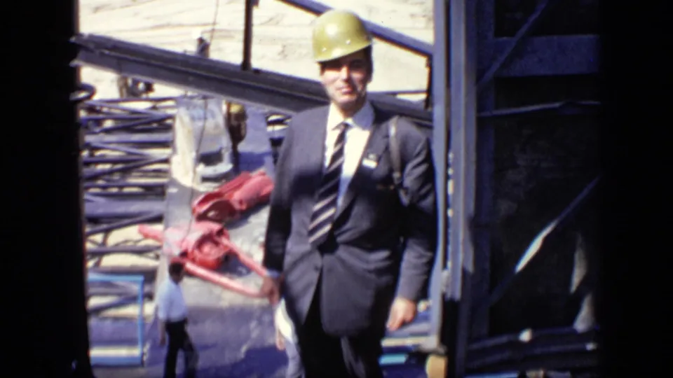 A suited visitor wearing a yellow hard hat walks toward the camera at a 1971 Moscow construction site with steel beams behind him.