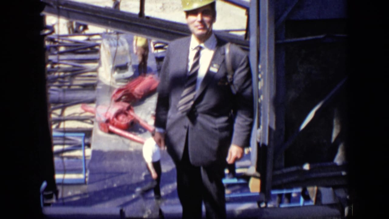 In Moscow in 1971, a suited man wearing a yellow hard hat stands on a construction site with machinery and water behind him.