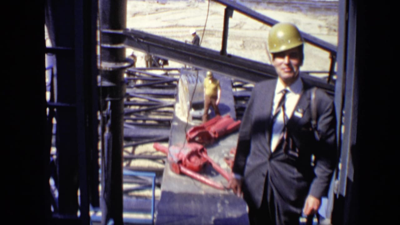 Suited man in yellow hard hat standing beside steel framing and red machinery.