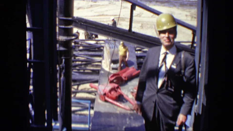 A suited man in a yellow hard hat stands near steel construction framing, with a worker and waterfront ground visible in the background.