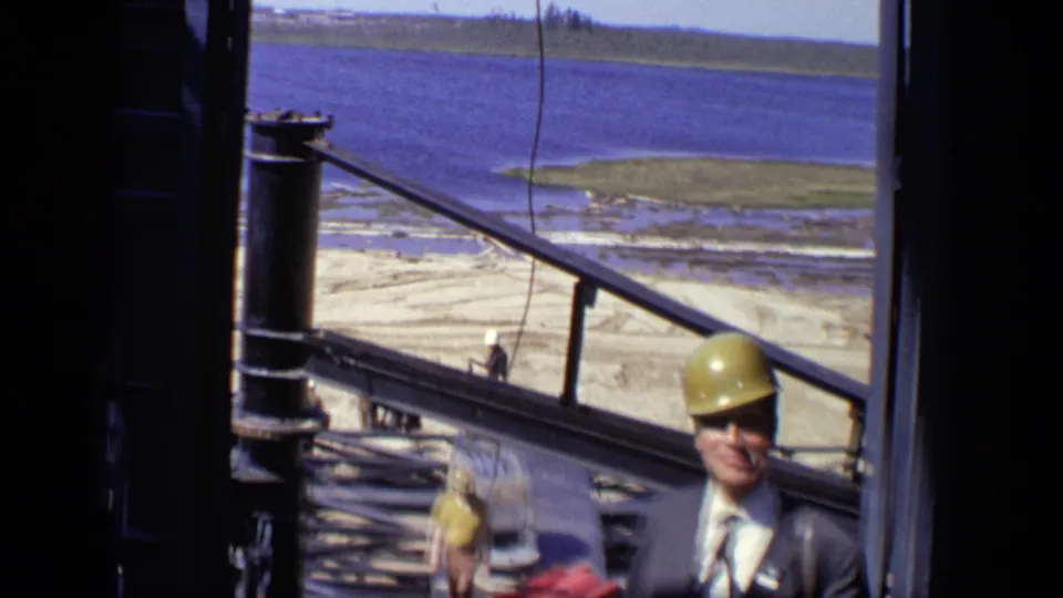 The frame looks across a waterfront construction site in Moscow, with metal railings, workers, and the suited hard-hat wearer partly visible.