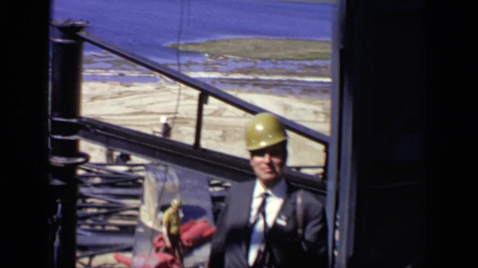 A suited man in a yellow hard hat stands by red machinery and metal framing at a Moscow construction site, with water visible beyond.
