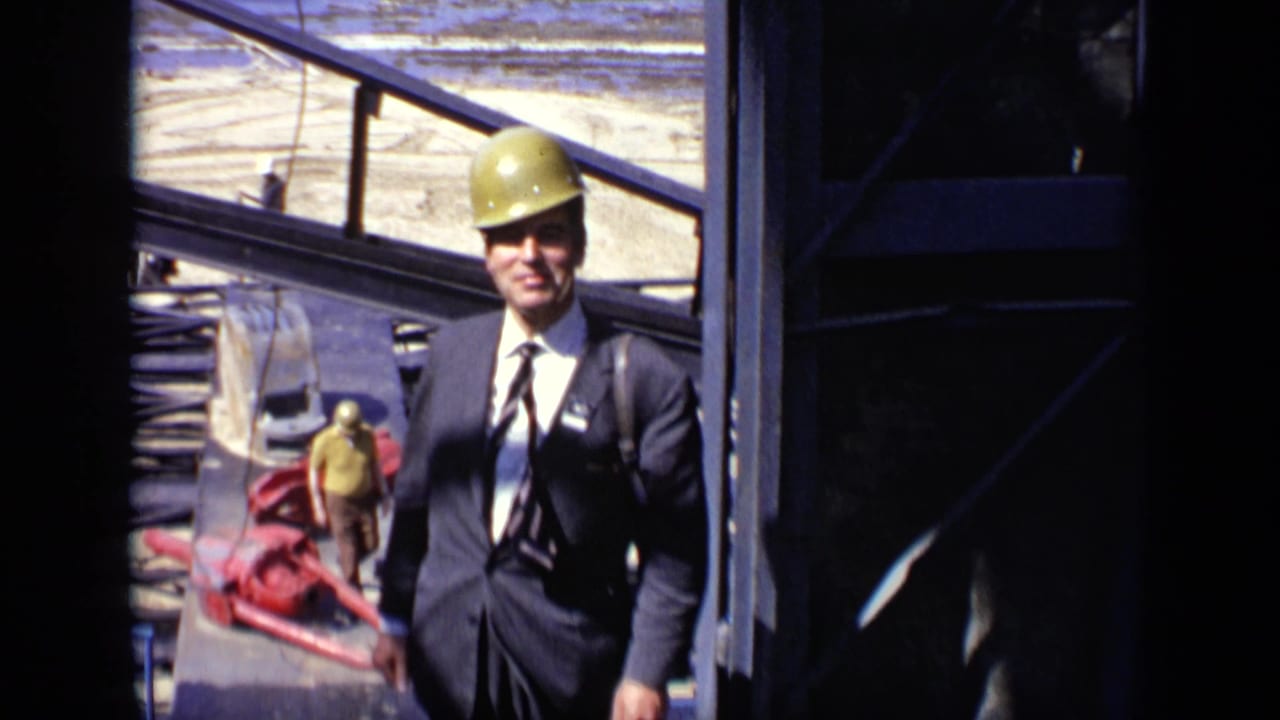 In 1971 Moscow footage, a suited man wearing a hard hat smiles near steel framing and construction materials.