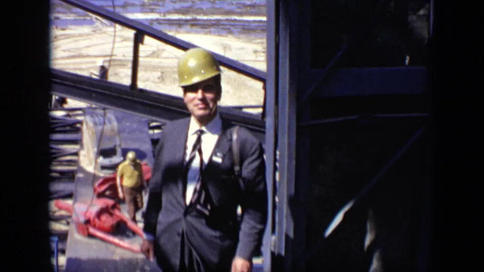 In 1971 Moscow footage, a suited man wearing a hard hat smiles near steel framing and construction materials.