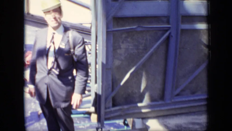 A close, handheld frame shows the suited man passing a metal-framed wall or site structure in the Moscow construction footage.
