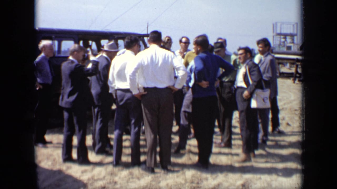 In 1971 Moscow, adult men in shirts, suits, hats, and sunglasses gather outdoors on a sunny dirt field near a van.