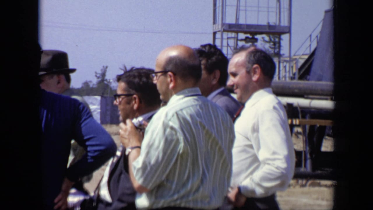 In 1971 Moscow home-movie footage, several adult men in shirts, suits, sunglasses, and a hat stand together near metal scaffolding.