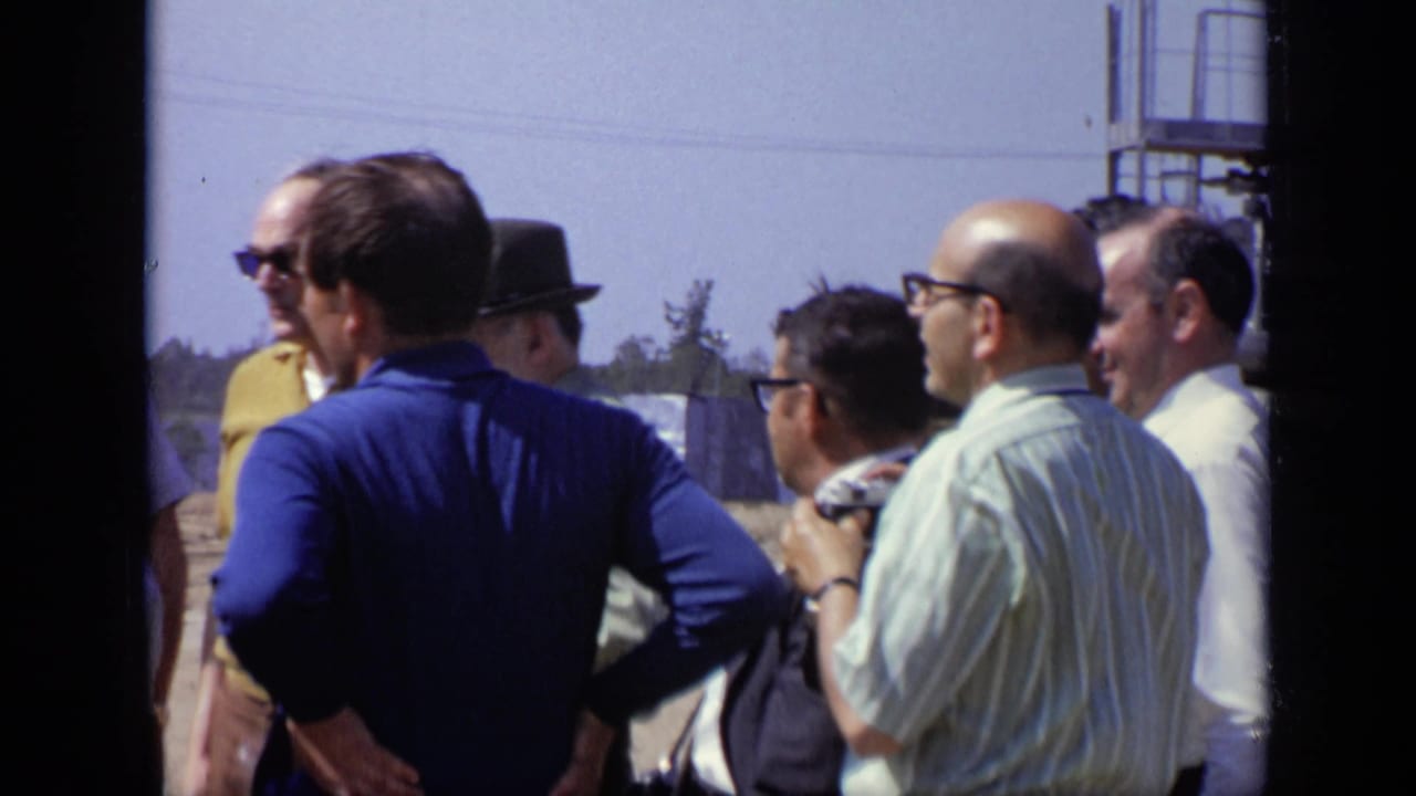 Several adult men in casual and formal shirts stand in a loose circle outdoors in Moscow, with industrial structures visible behind them.