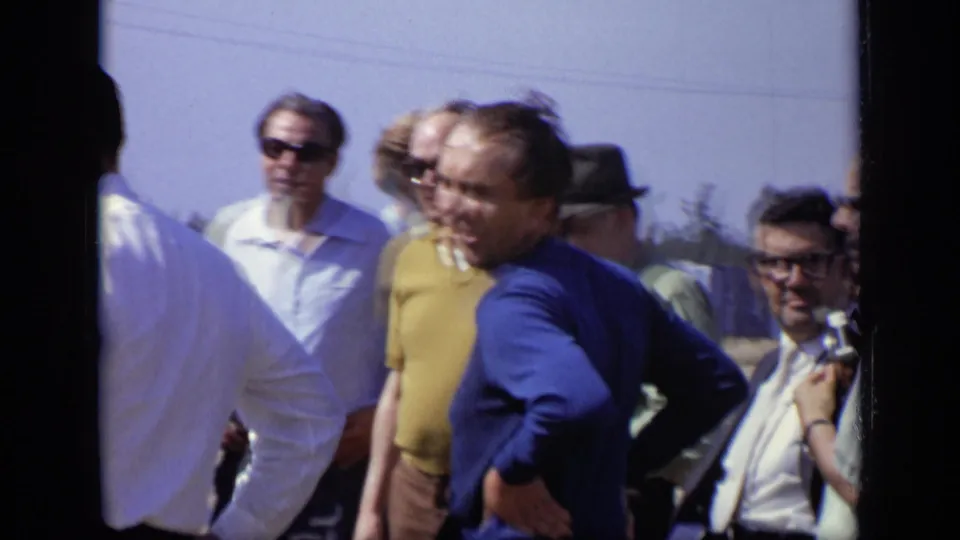 A handheld 8mm frame shows adult men outdoors in Moscow, including a man in a blue sweater and another in a yellow shirt near the group.