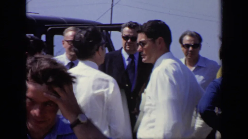 Adult men in shirts, suits, and sunglasses stand beside a dark vehicle in 1971 Moscow home-movie footage, with faces visible.