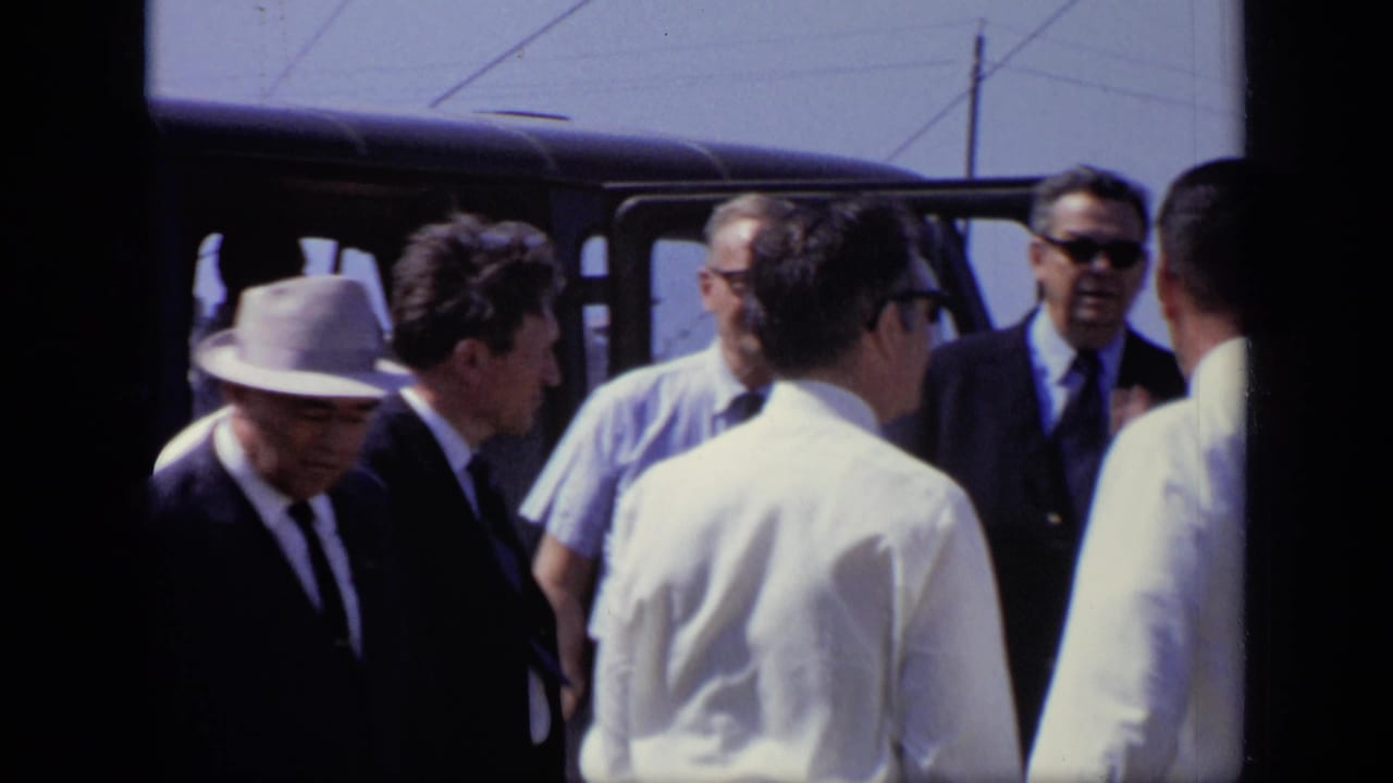 1971 Moscow home-movie frame showing suited men and workers gathered near a dark van at a construction site.