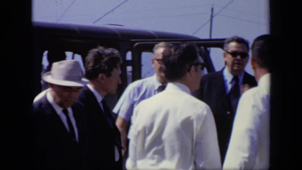 1971 Moscow home-movie frame showing suited men and workers gathered near a dark van at a construction site.