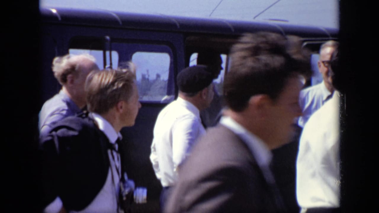 A group of men stands near a dark van, with one man passing close to camera.