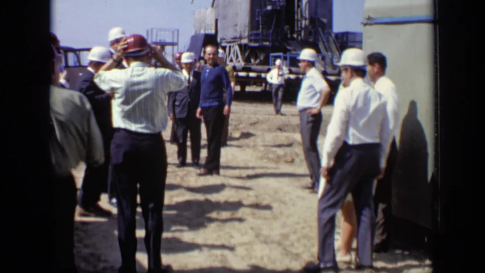 Wide construction-site view in Moscow, 1971, with hard-hatted men, suited visitors, and industrial equipment in the background.