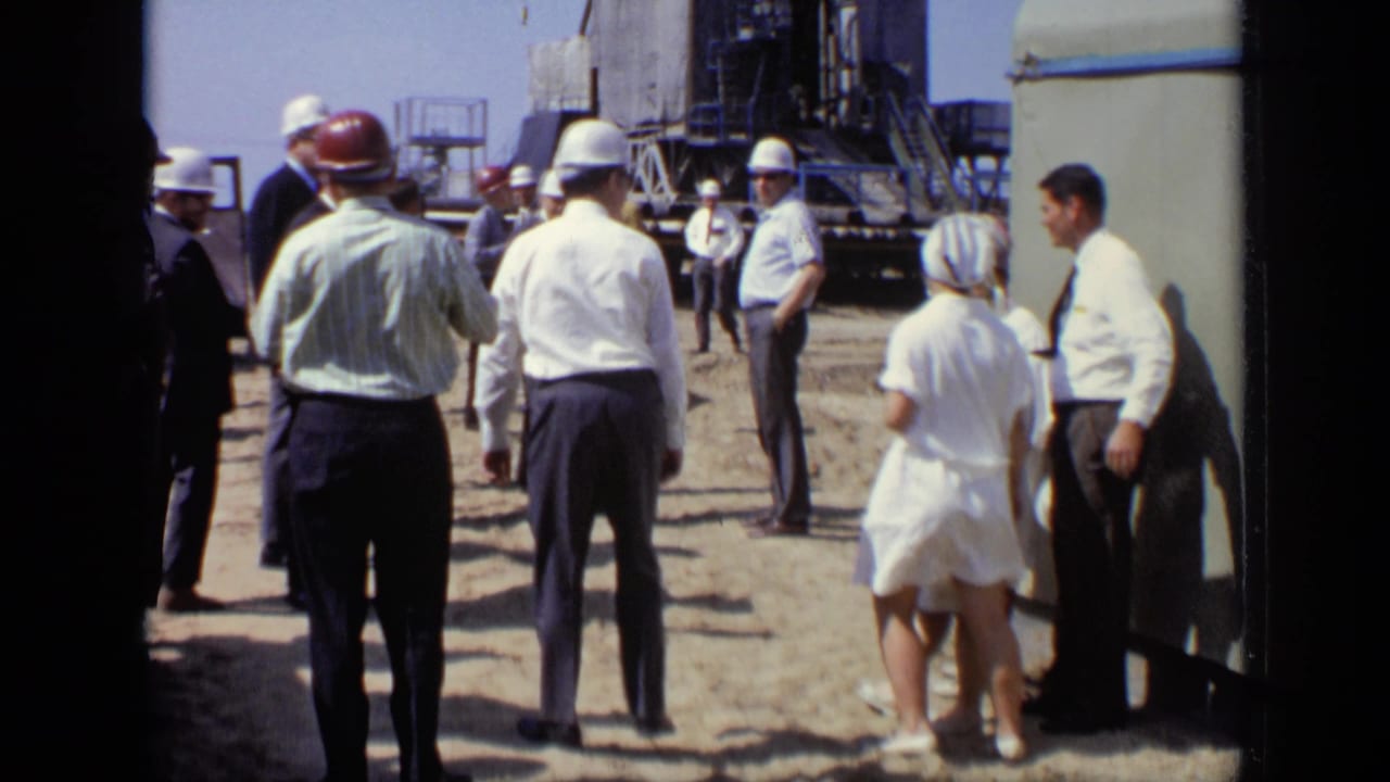 Adults in hard hats and summer clothing stand near a large industrial structure.