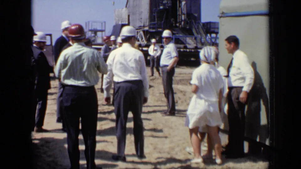 1971 Moscow home-movie frame showing adults in hard hats, suits, and white clothing gathered at an industrial site.