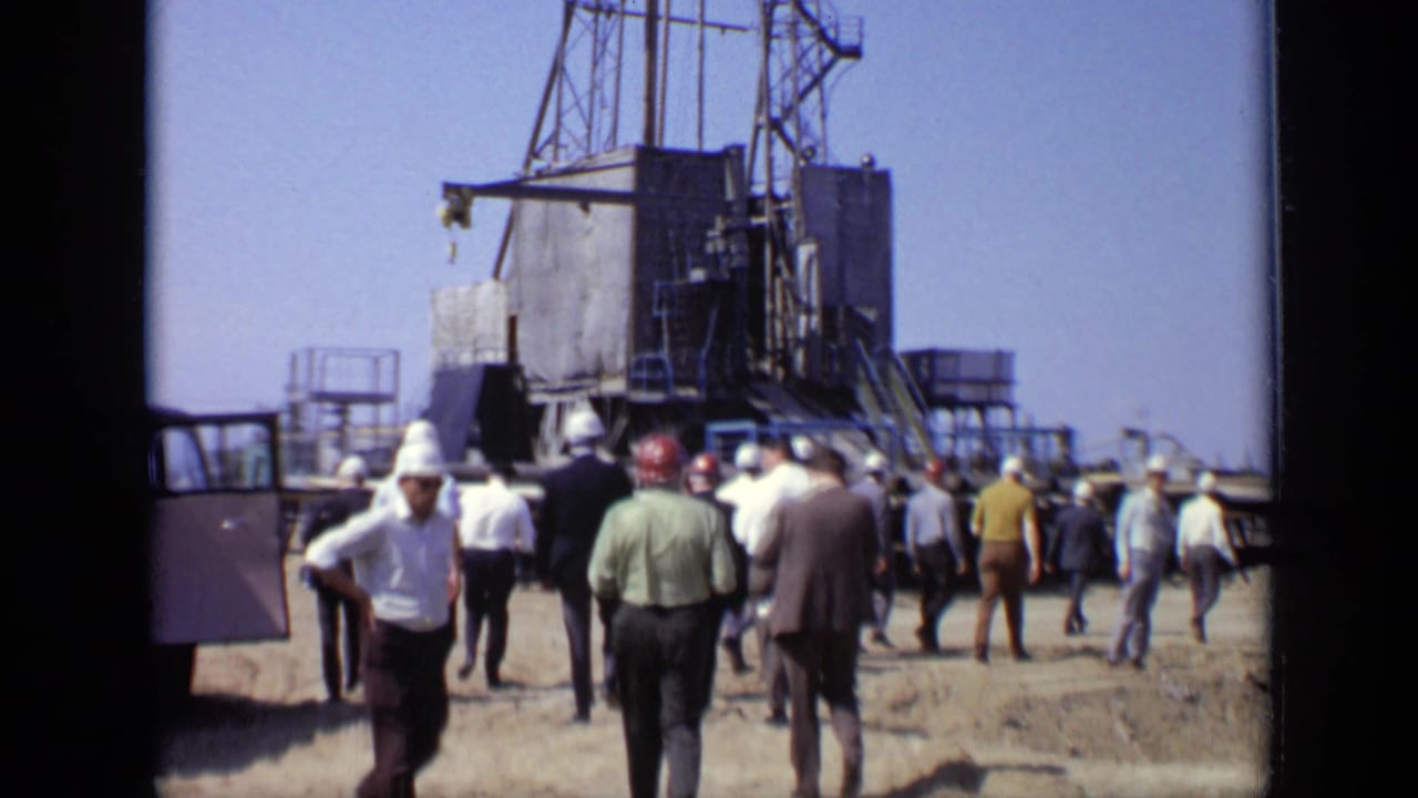 1971 Moscow 8mm frame showing workers in hard hats and men in period clothing approaching a tall metal rig at an industrial site.