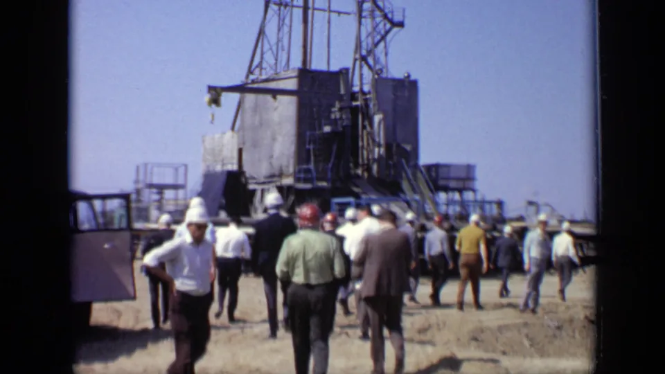 1971 Moscow 8mm frame showing workers in hard hats and men in period clothing approaching a tall metal rig at an industrial site.