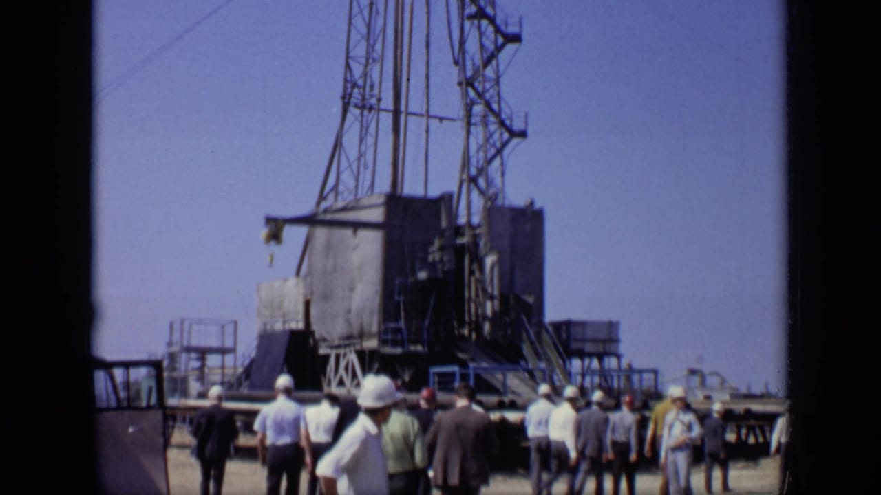 1971 Moscow home-movie frame with workers and observers clustered near a metal lattice rig at an outdoor industrial site.