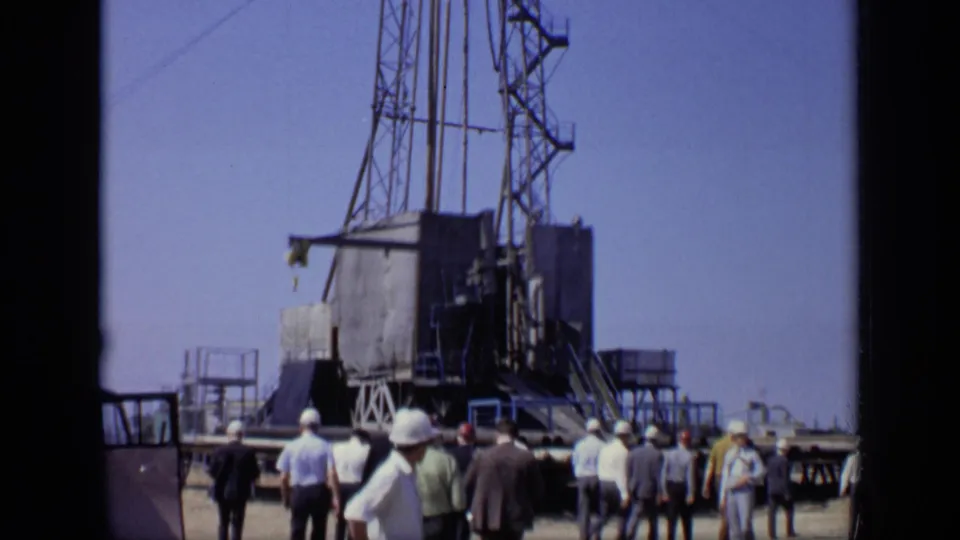 1971 Moscow home-movie frame with workers and observers clustered near a metal lattice rig at an outdoor industrial site.