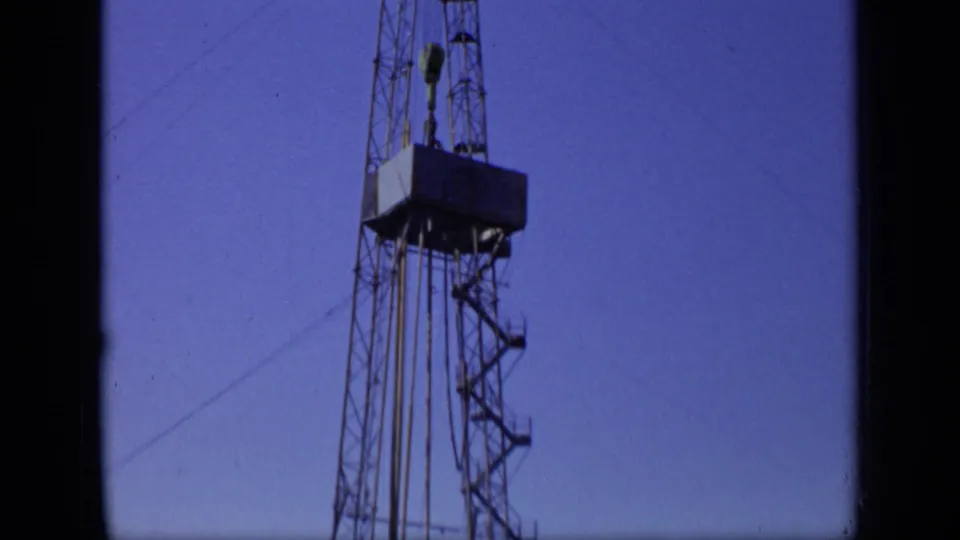 1971 Moscow home-movie frame showing a tall lattice oil rig tower with a platform and cables against a deep blue sky.