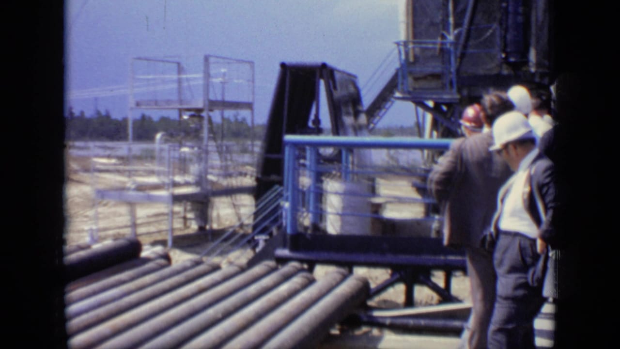 1971 Moscow home-movie frame showing suited adults and hard-hatted personnel near large pipes, blue railings, and industrial structures.