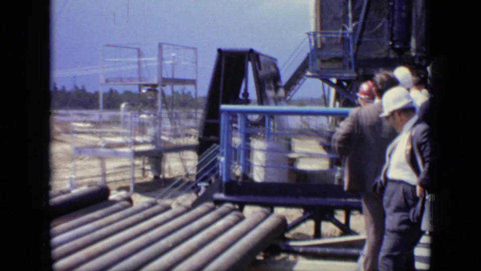 1971 Moscow home-movie frame showing suited adults and hard-hatted personnel near large pipes, blue railings, and industrial structures.