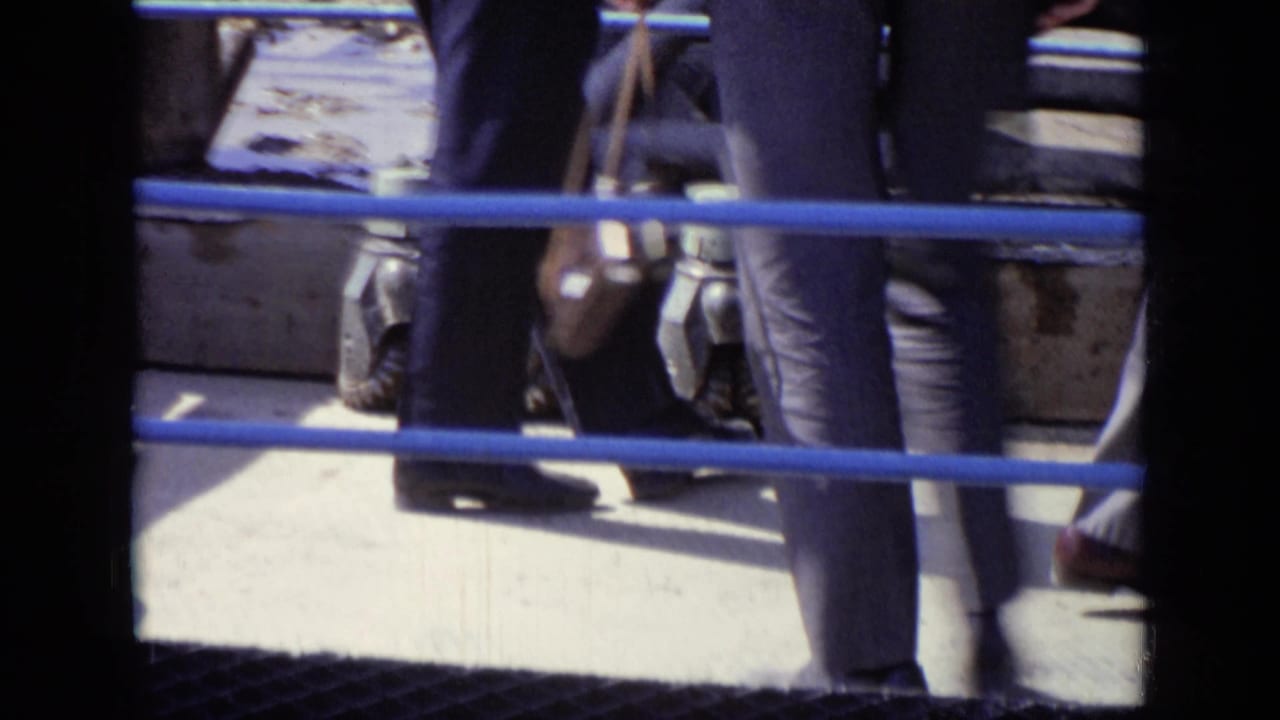 Lower bodies of men walking behind blue railings beside industrial equipment.