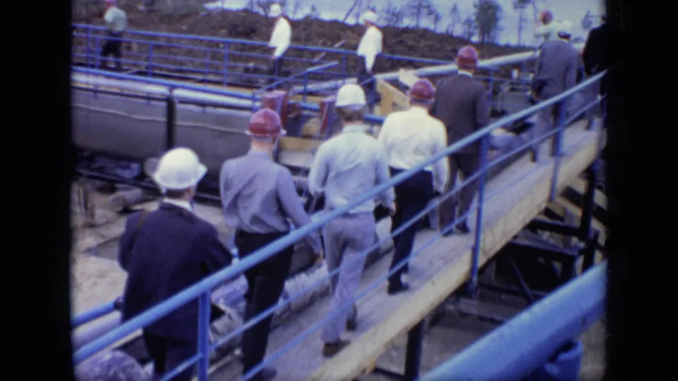 A group of workers and visitors in hard hats walk along a wooden walkway at an outdoor Moscow industrial site in 1971.