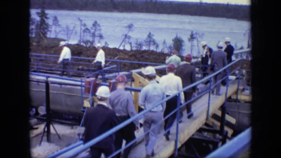 Workers and visitors continue along a blue-railed walkway, with water and a tree-lined shore beyond the 1971 Moscow industrial site.