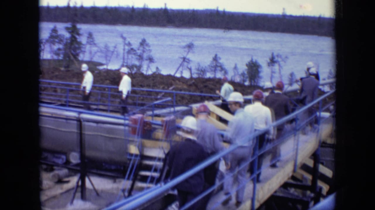 1971 Moscow home-movie frame of hard-hatted workers and suited men on a blue-railed riverside industrial platform.