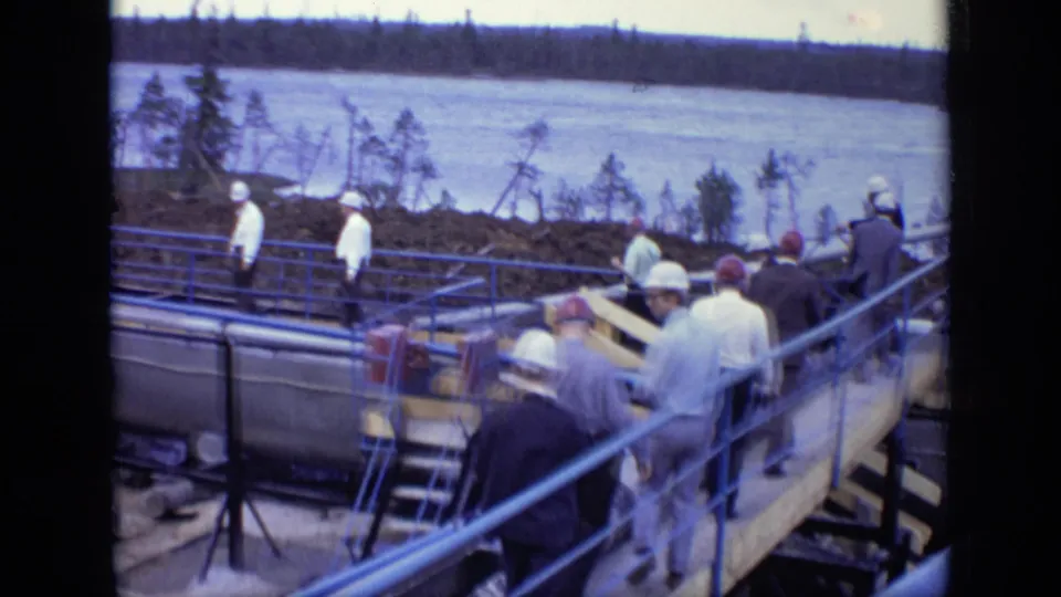 1971 Moscow home-movie frame of hard-hatted workers and suited men on a blue-railed riverside industrial platform.