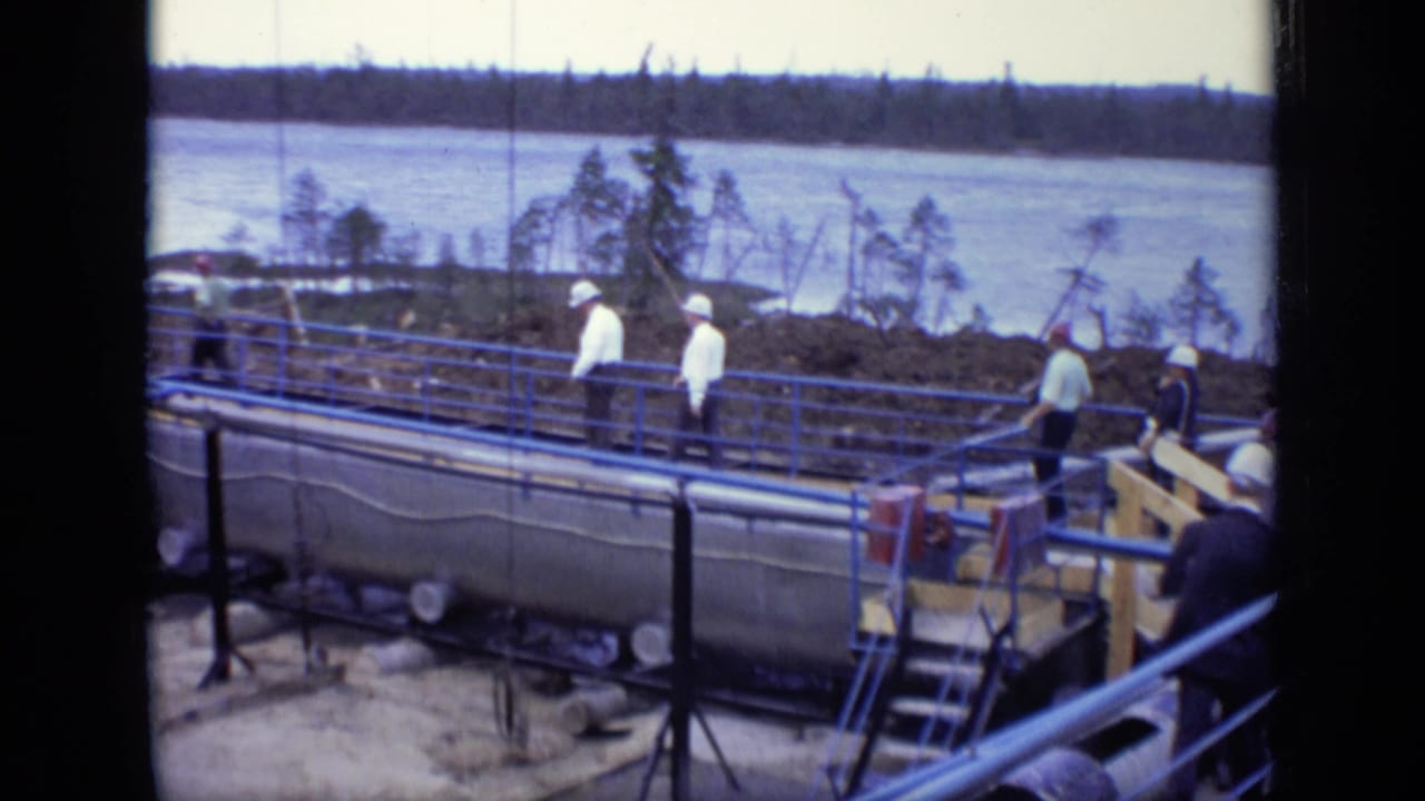 Several men walk on a blue-railed industrial walkway with tanks and a river in the background.