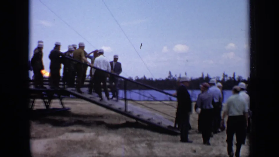 1971 Moscow home-movie frame of men in hard hats and suits gathered near a ramp at a riverside worksite.