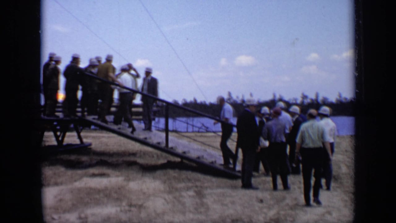Men in hard hats and suits gather near a metal ramp beside water.