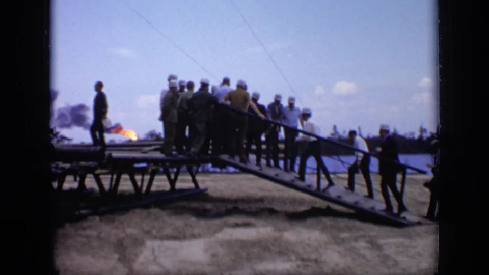 1971 Moscow industrial visit frame with men on a metal ramp and distant smoke or flame at the left edge of the site.