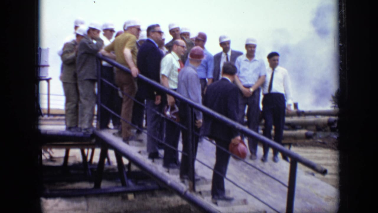 In 1971 Moscow, a group of men in hard hats, suits, and work attire stand on a ramp near pipes and smoke at an industrial site.