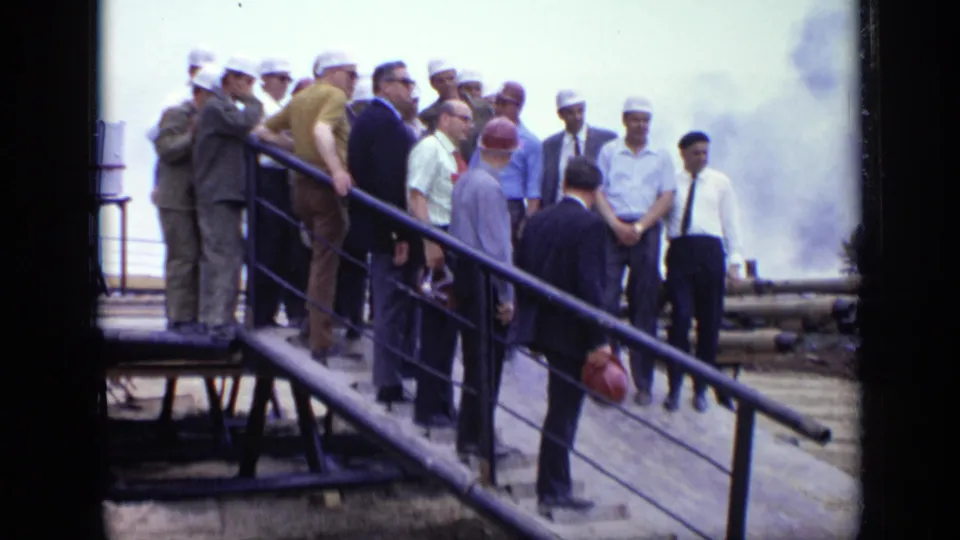 In 1971 Moscow, a group of men in hard hats, suits, and work attire stand on a ramp near pipes and smoke at an industrial site.