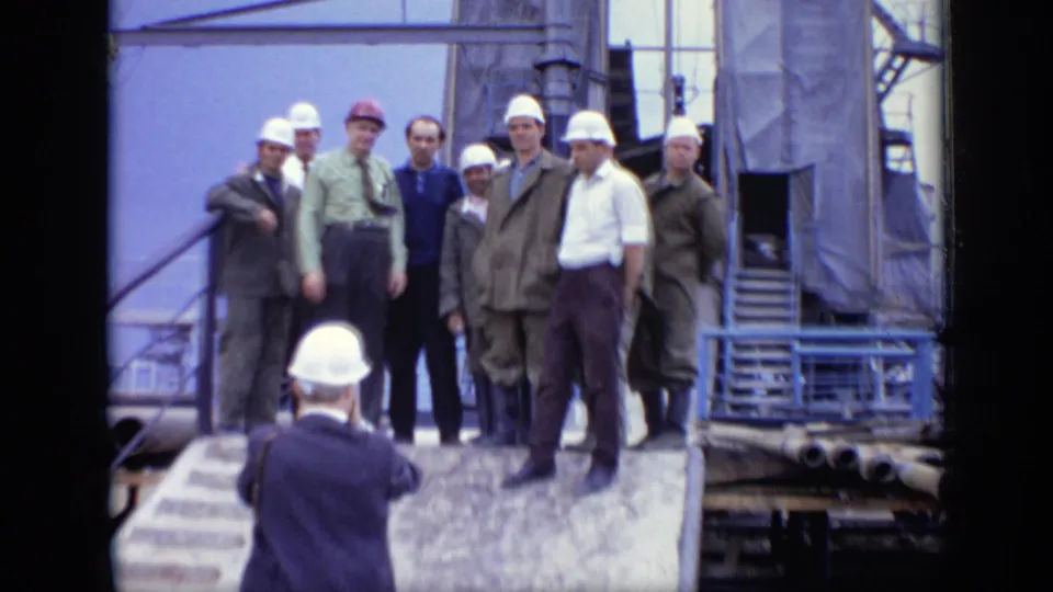 At the Moscow industrial site, a hard-hatted figure in the foreground faces a posed group of workers and officials on a ramp near scaffolding.